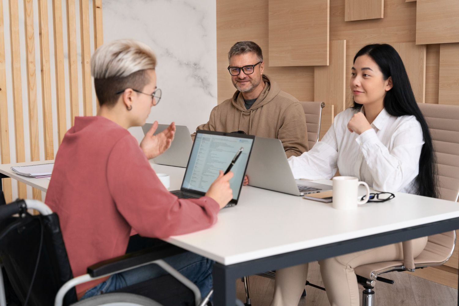 Person in wheelchair, pointing to a laptop screen, discussing with two colleagues at a table in an office setting.