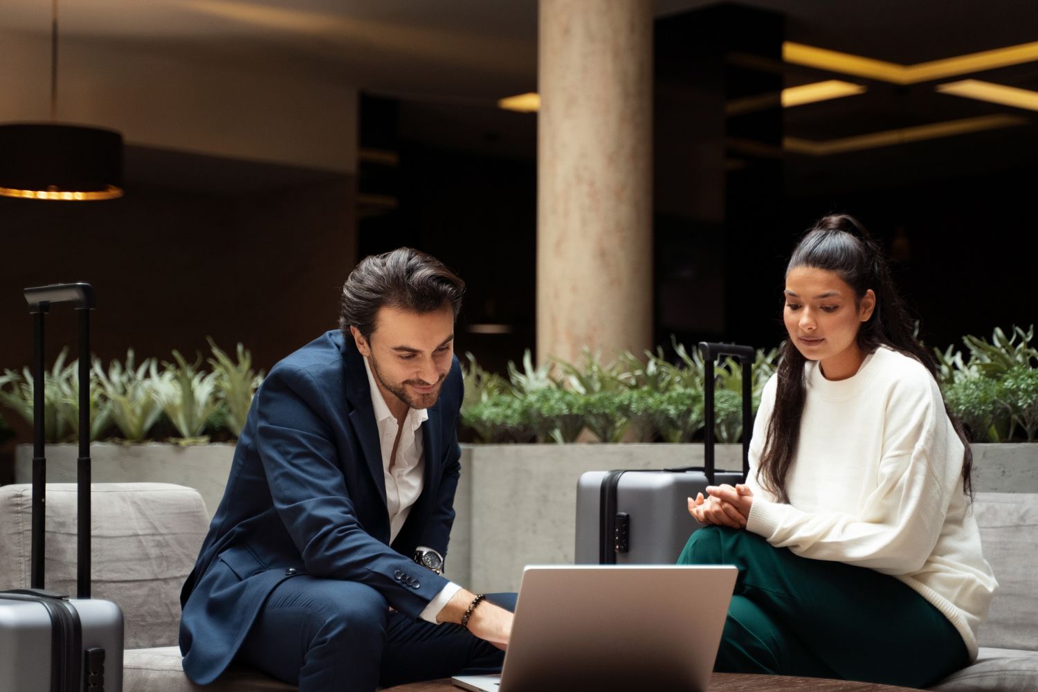 A man and woman sit, looking at a laptop in a hotel lobby, luggage nearby.