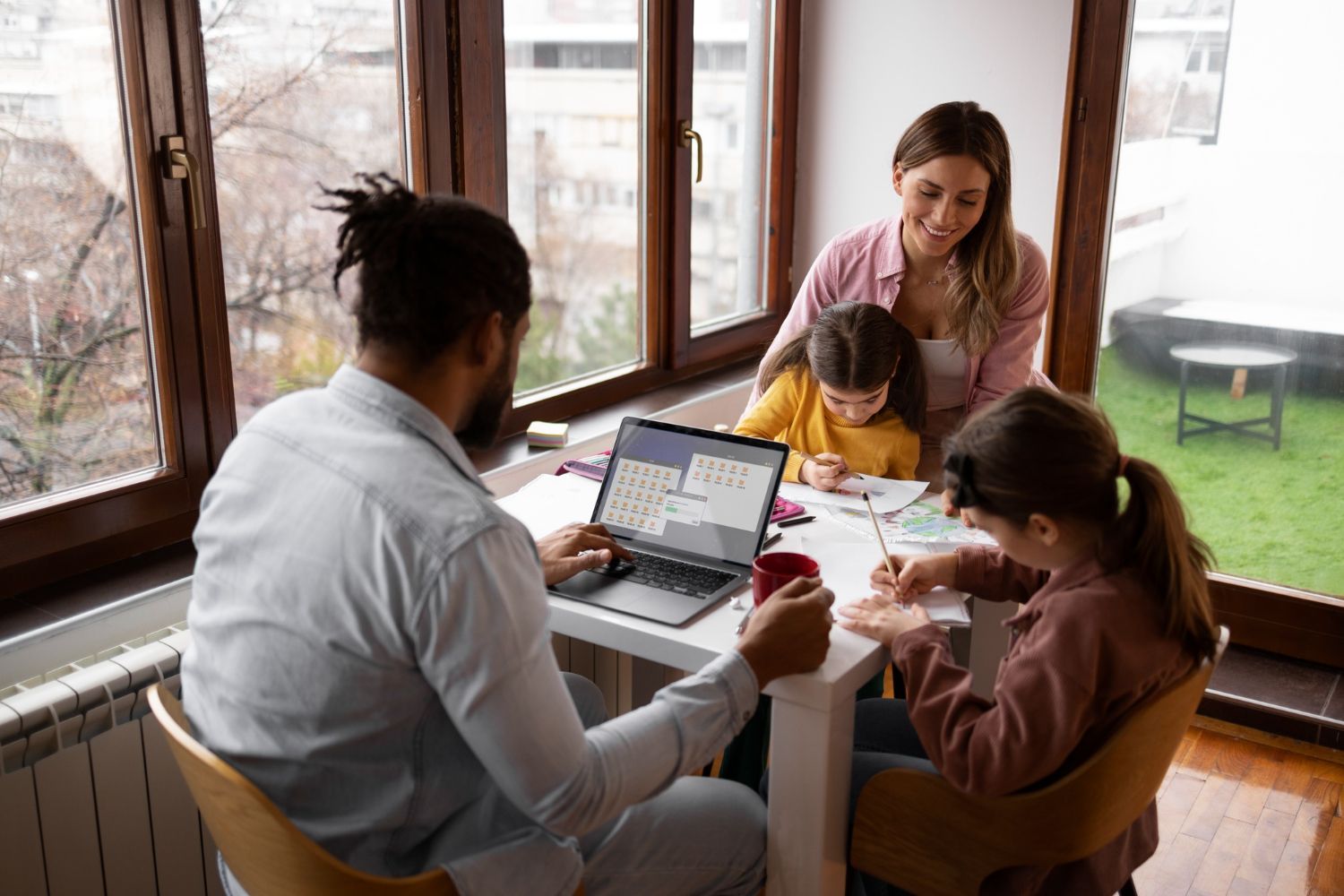 Family working at a table: man on laptop, two children drawing, woman watching. Near a window.