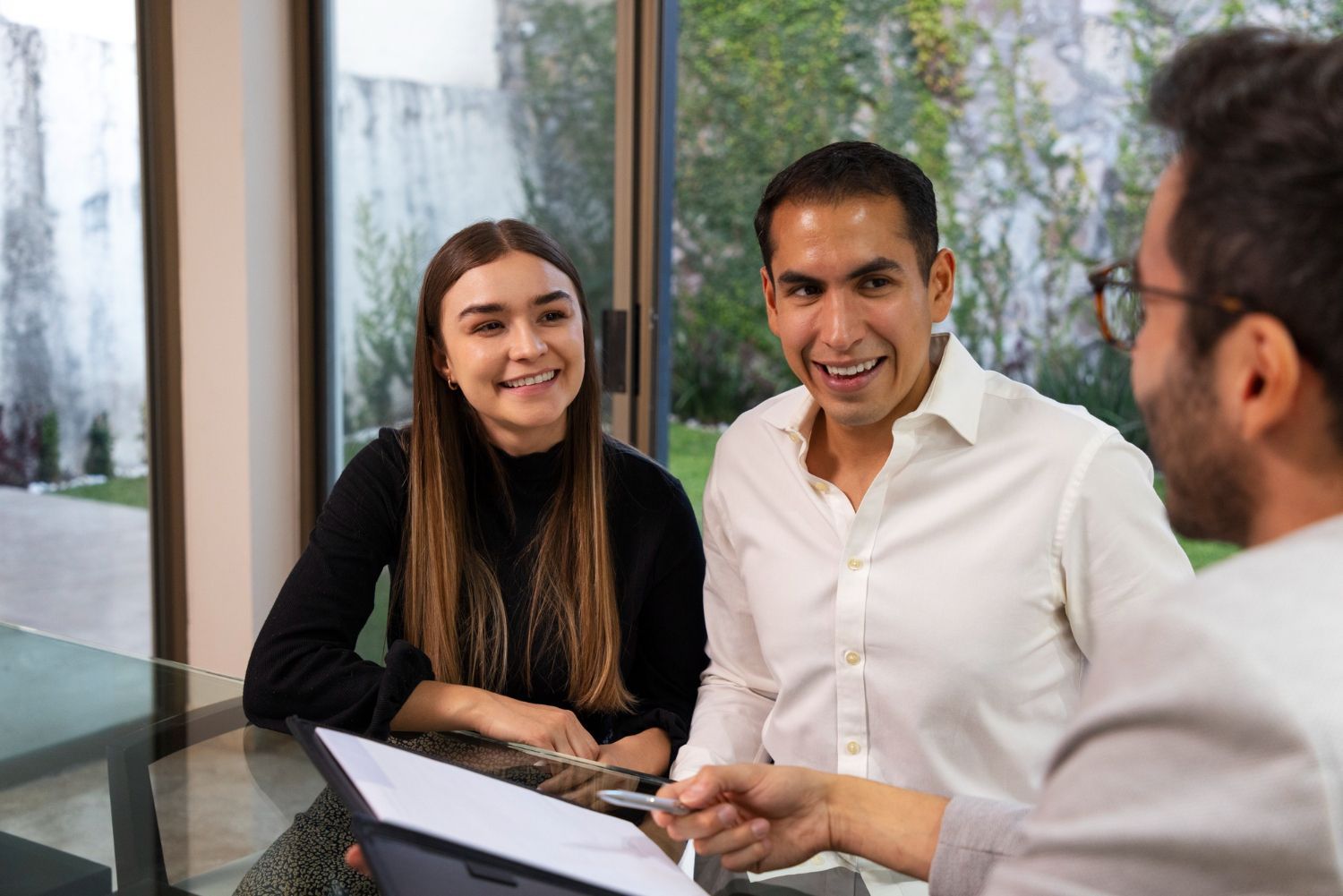 A couple smiles while reviewing documents with a professional, seated at a table near a window.
