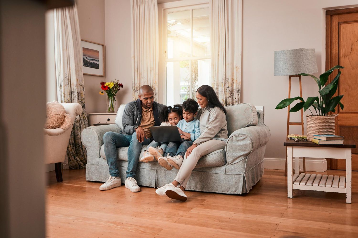 Family of four sitting on couch, looking at tablet in bright living room.