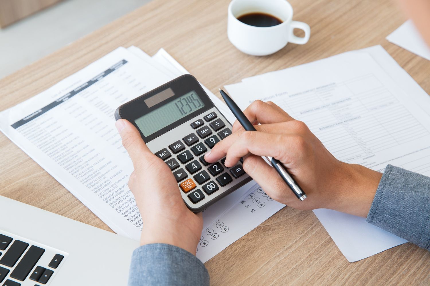 Hands using a calculator on a desk with paperwork and a coffee cup.