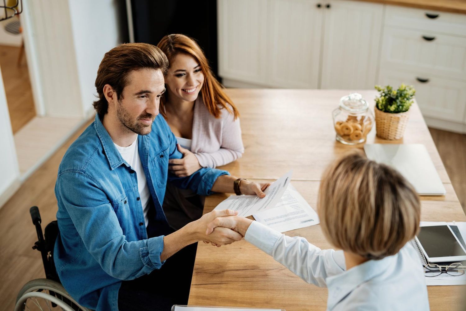 Couple with a wheelchair user shakes hands with a person at a table, likely finalizing documents in a home.