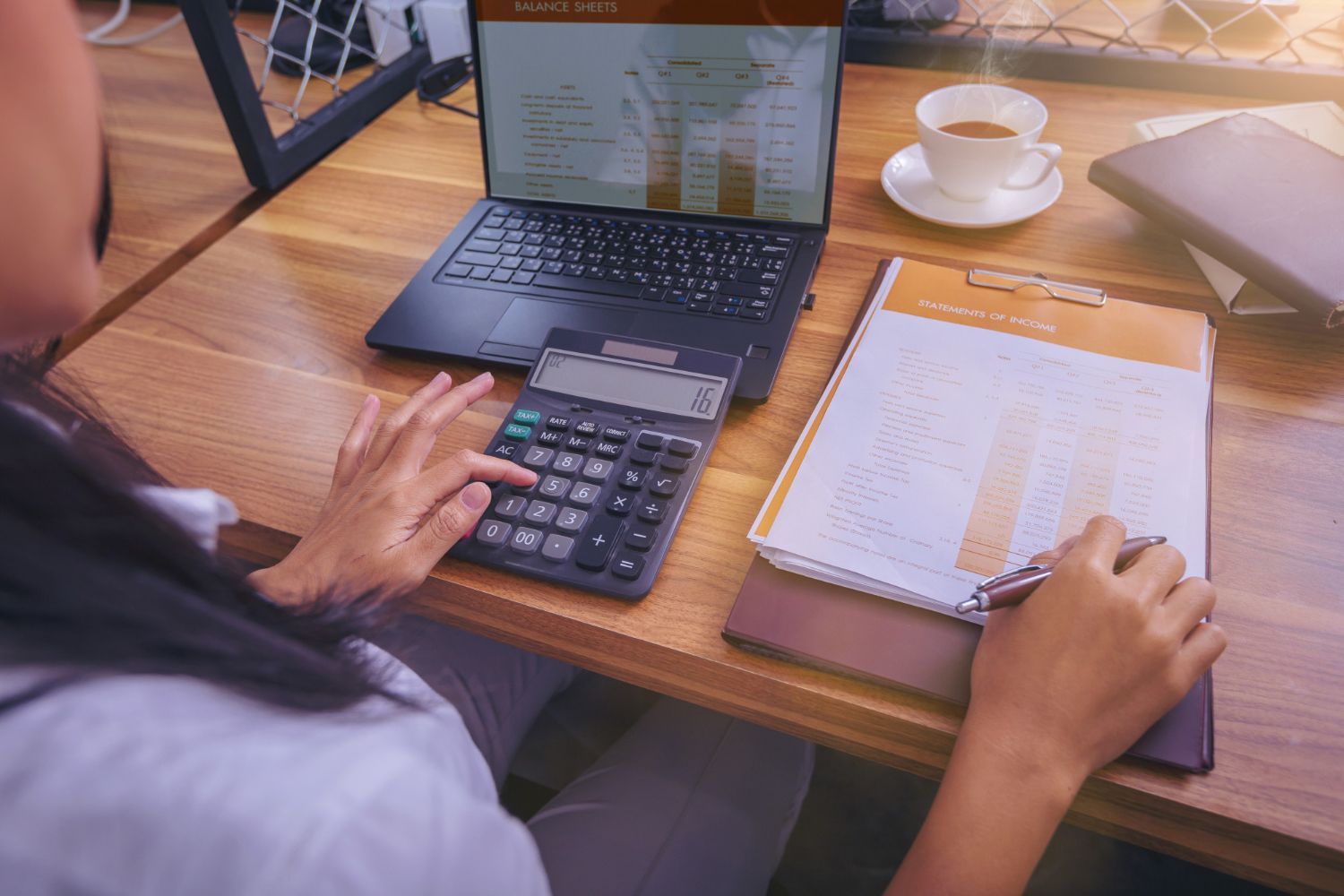 Person using a calculator, laptop displaying charts, paperwork, and coffee on a wooden desk.