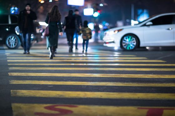 People crossing a street at night using a crosswalk. A white car is approaching.