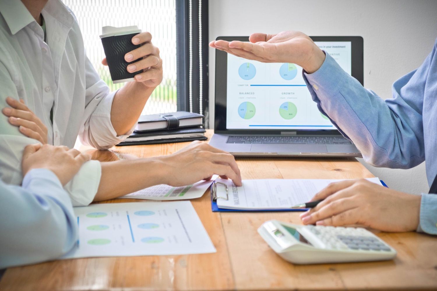 People in office meeting, reviewing graphs and documents. One gesturing, another holding coffee.