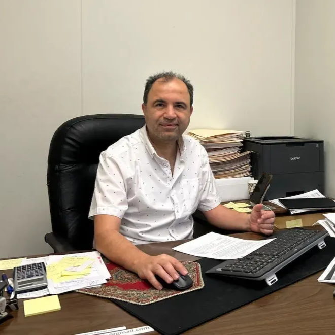 Man in white shirt sits at a desk with computer and documents; flags of Afghanistan and Poland in the background.