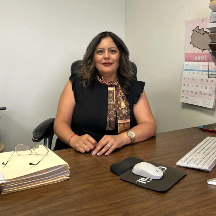 Woman seated at a desk, looking at the camera. She's in an office setting with a computer, and office supplies.