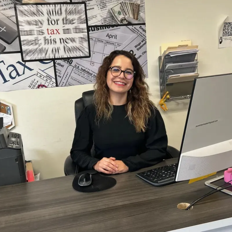 Woman at desk in office, smiling, with a computer and tax documents in the background.