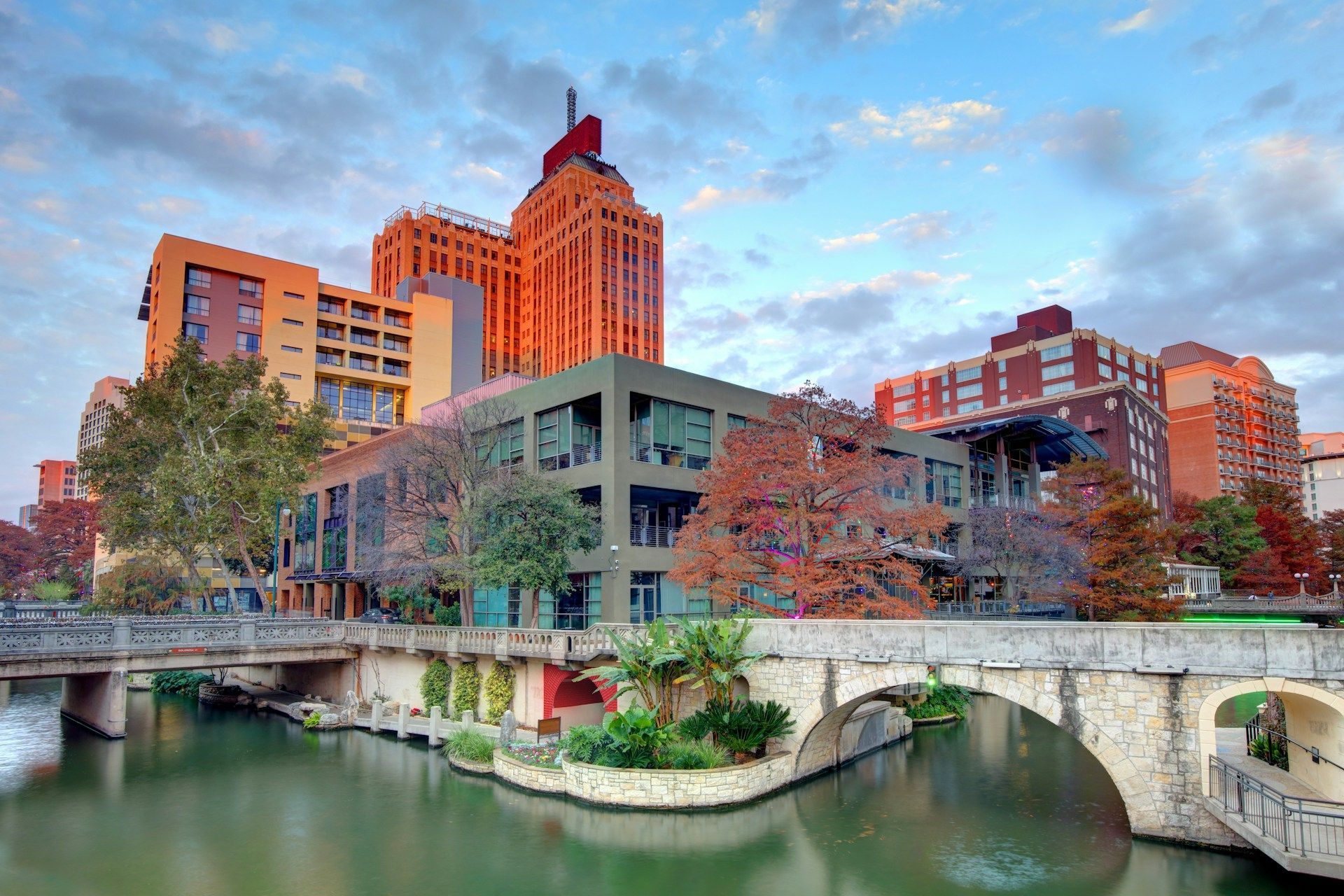 Riverwalk in San Antonio, Texas, with stone bridge, buildings, and water, under a cloudy sky.