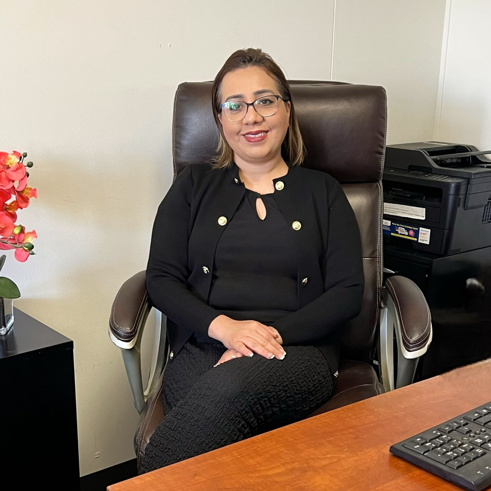 Woman seated at a desk in an office setting. Wearing a black suit, smiling. Desk with computer and printer.