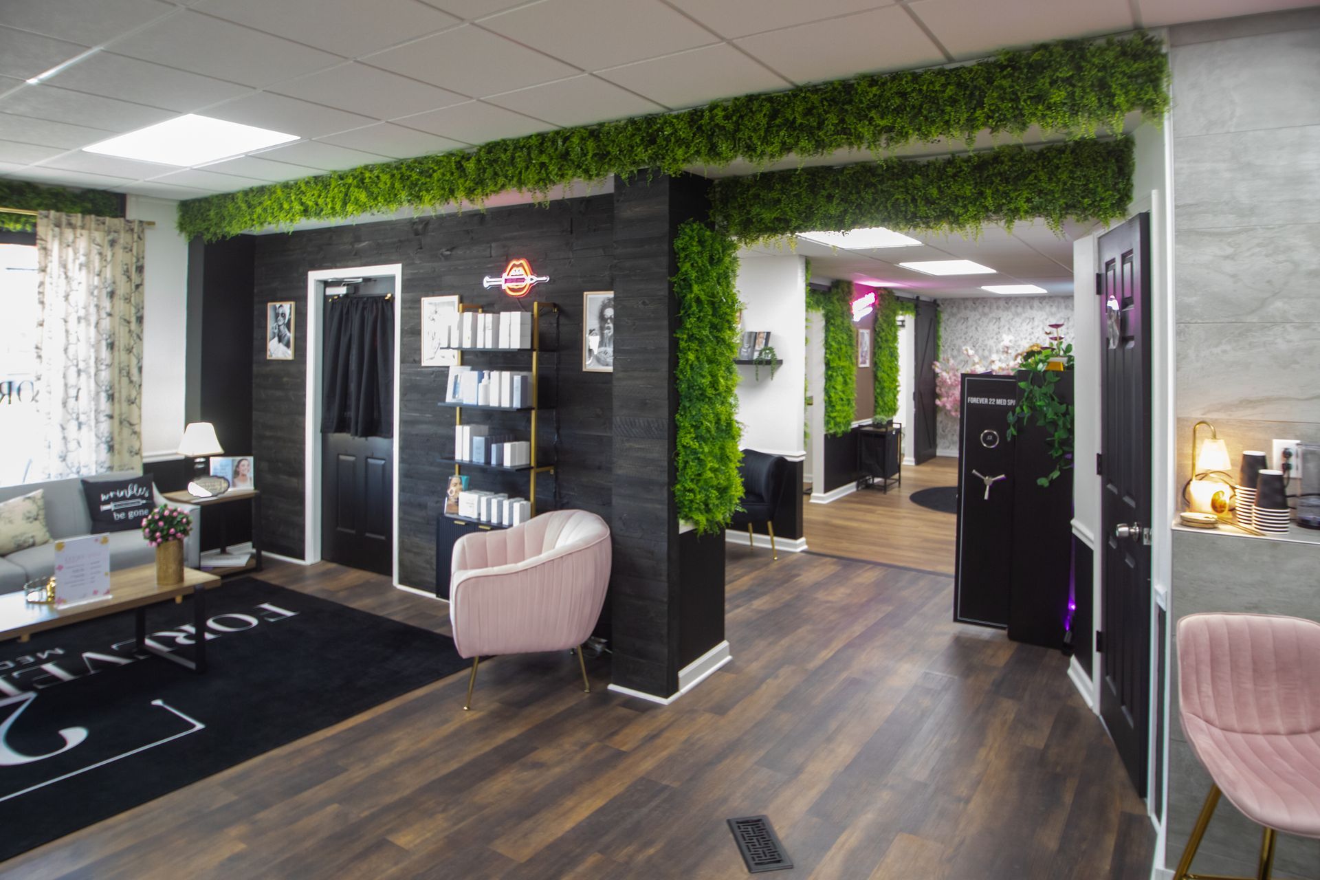 Two women conversing in a beauty salon reception area, with a desk, greenery wall, and a treatment room visible.