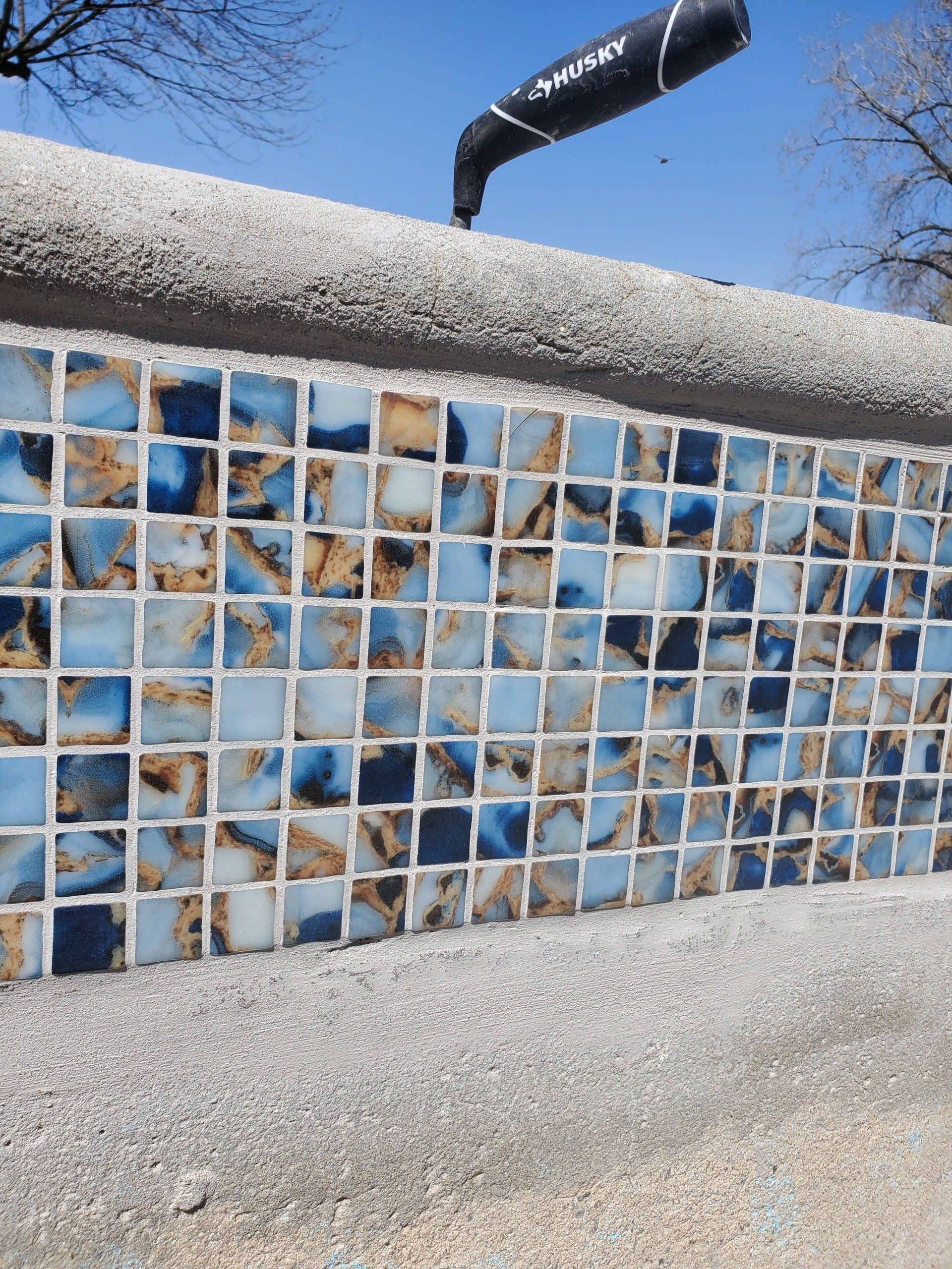 Carreaux de mosaïque bleus et marron sur un mur, surmontés d'une bordure grise, sur fond de ciel bleu.