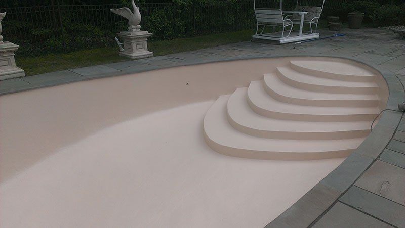 Piscine vide avec des marches incurvées et un intérieur beige, entourée d'une terrasse en pierre et de statues.
