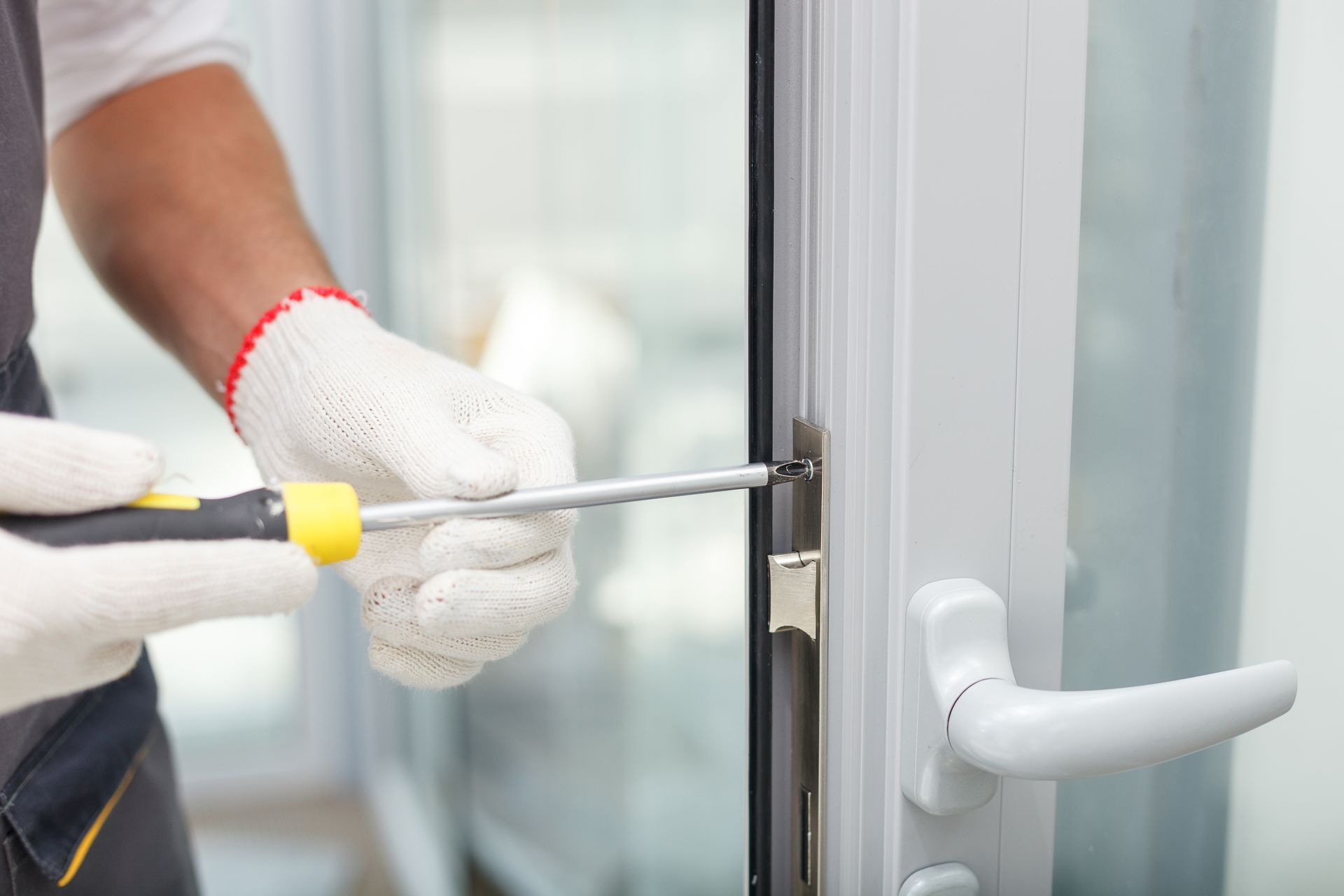 Gloved hands using a screwdriver to adjust a white door lock on a glass door
