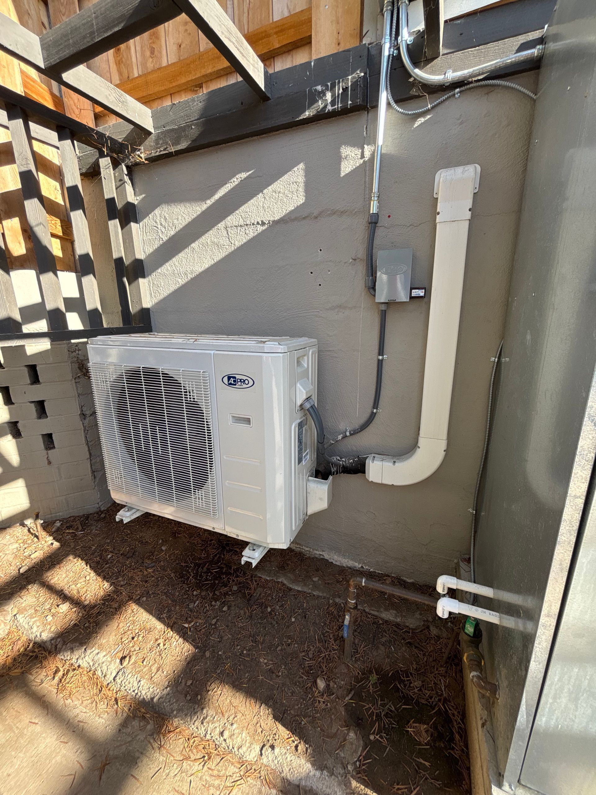 Exterior heat pump unit installed on a concrete wall with electrical conduit and drainage pipe.