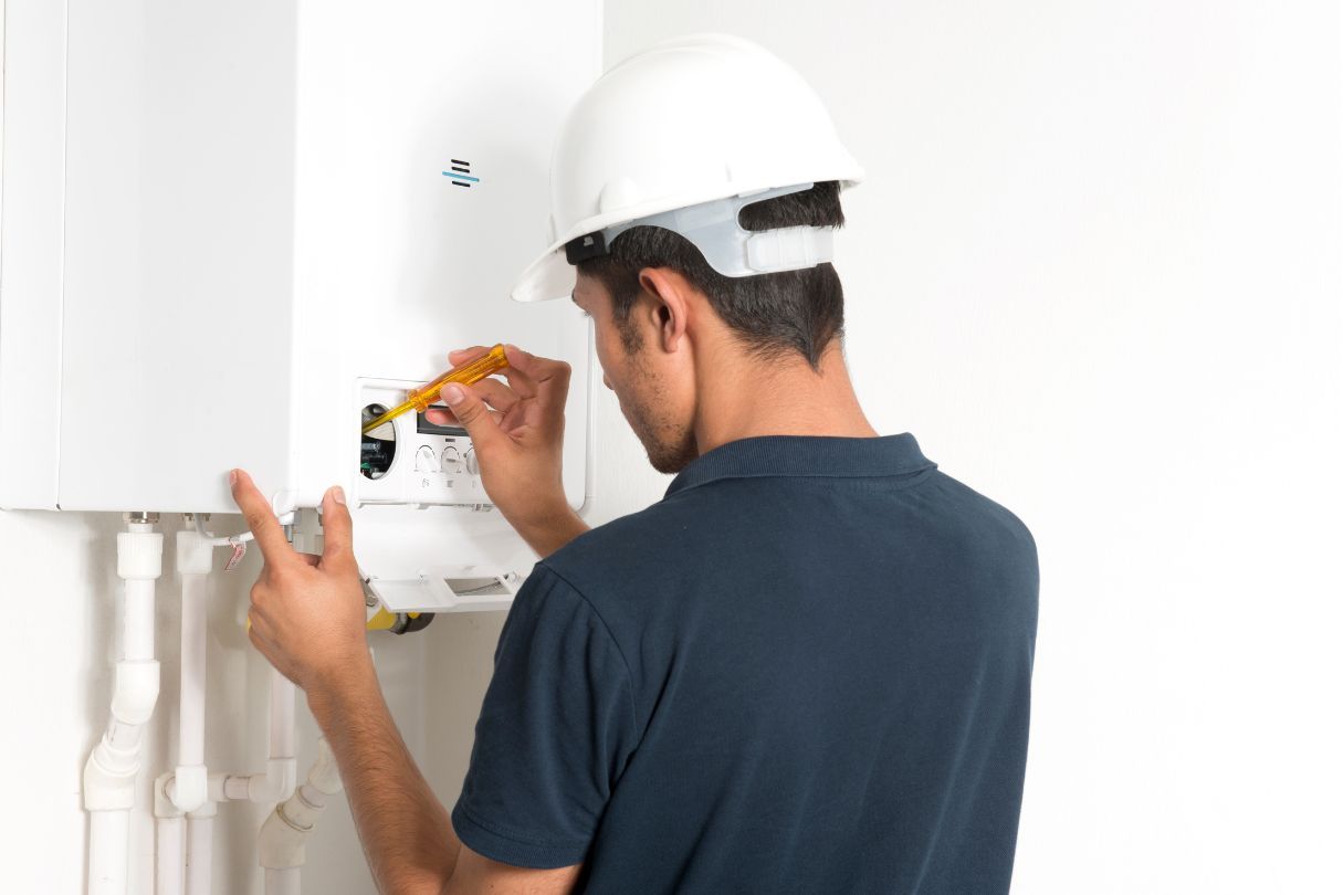 A man with a white hard hate, using a screwdriver on a white boiler connected to the wall