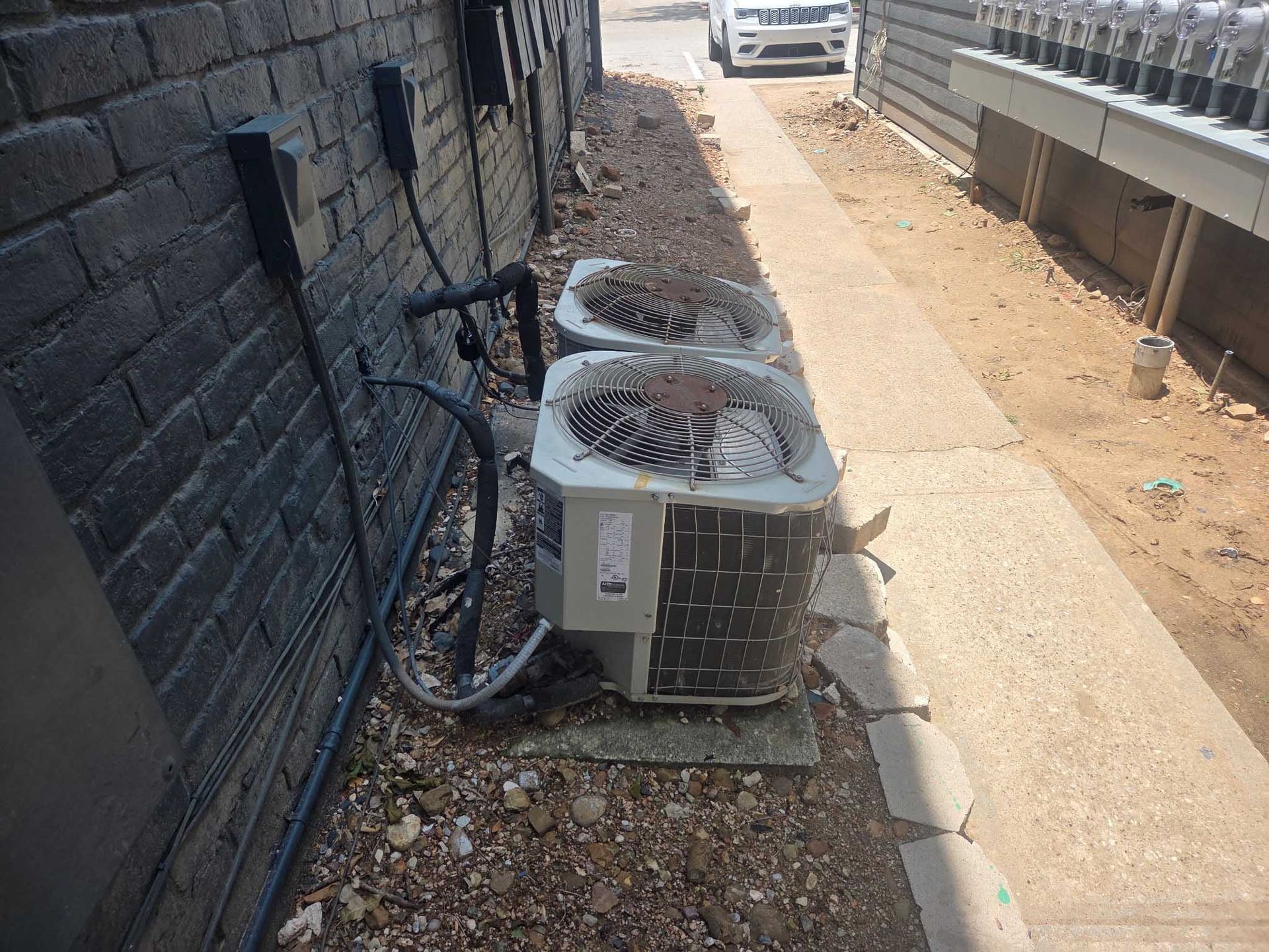 Two air conditioners are sitting on the sidewalk next to a brick wall.