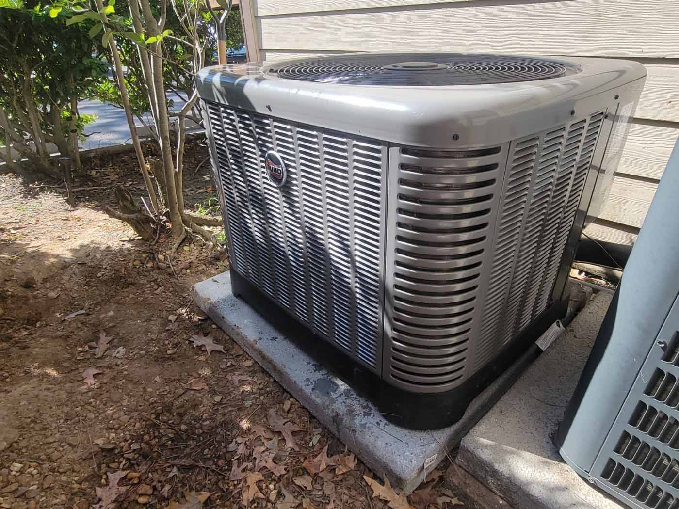 A large air conditioner is sitting on top of a concrete block next to a house.
