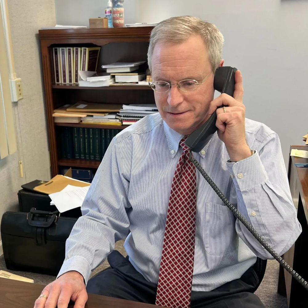  Brian Flanagan - Man in office, on phone, wearing glasses, striped shirt, and patterned tie. Bookshelf in background.
