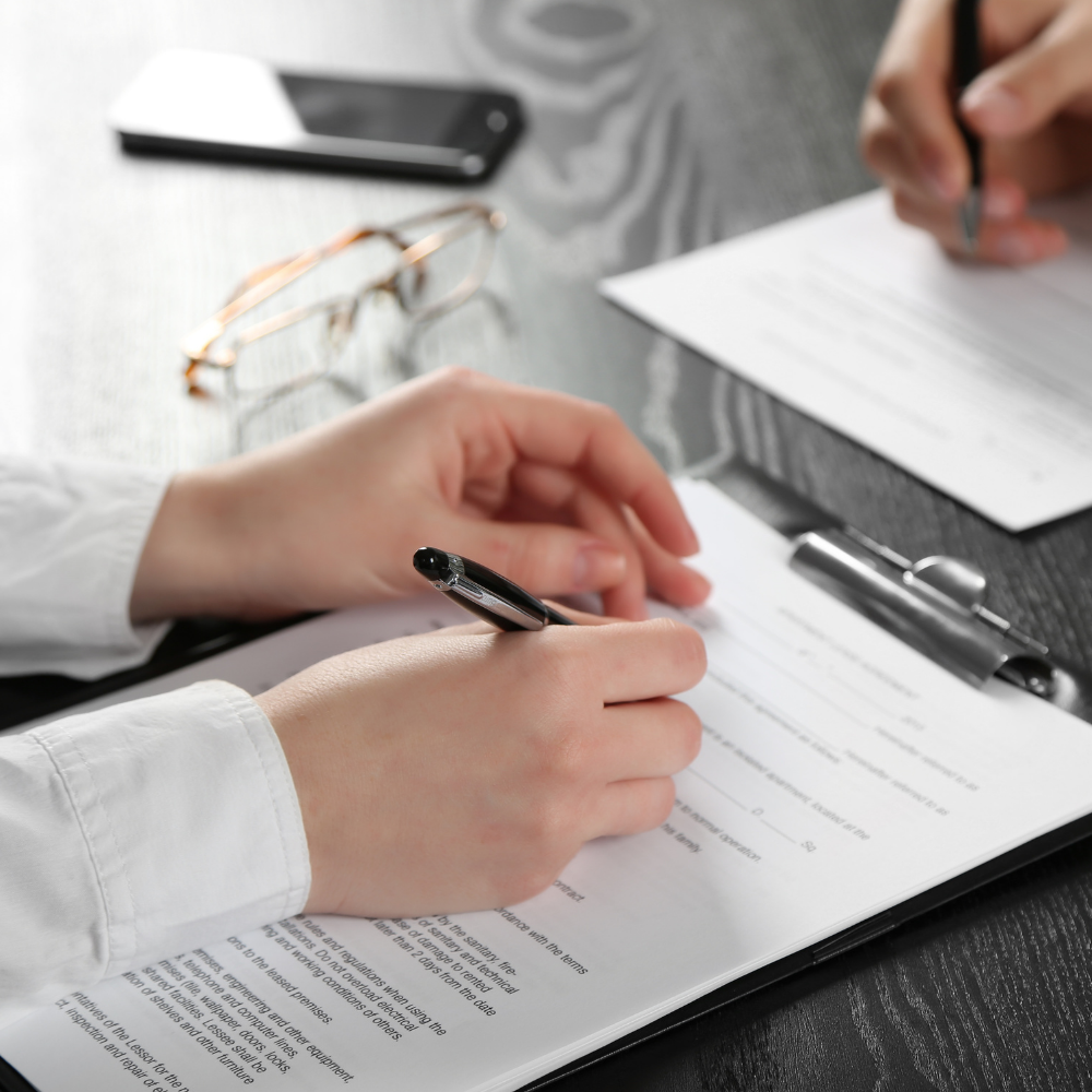 Hands signing documents on a clipboard and paper. A phone and glasses are on a table.