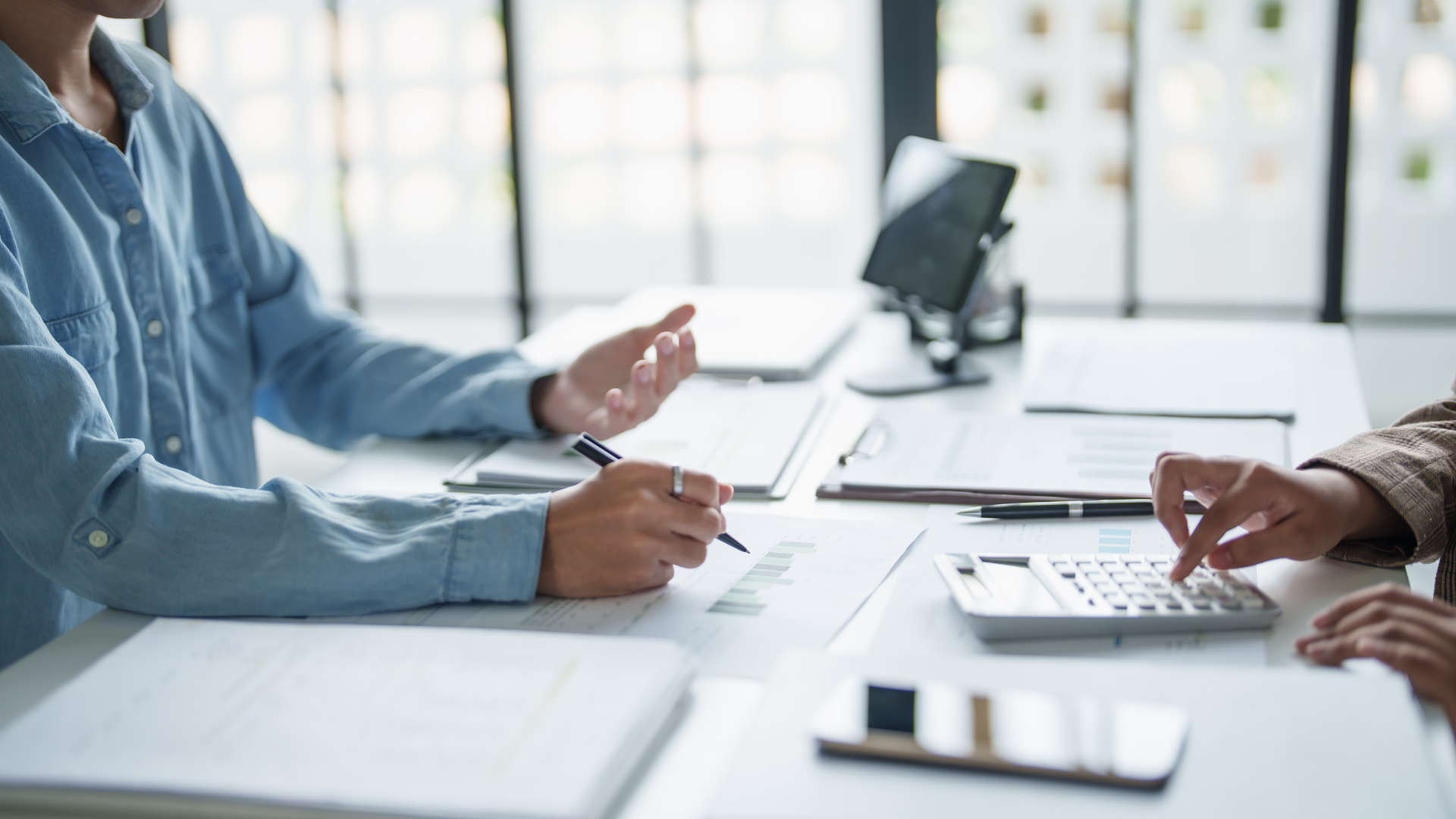 Two people in suits at a table reviewing documents, one gesturing towards the paper.