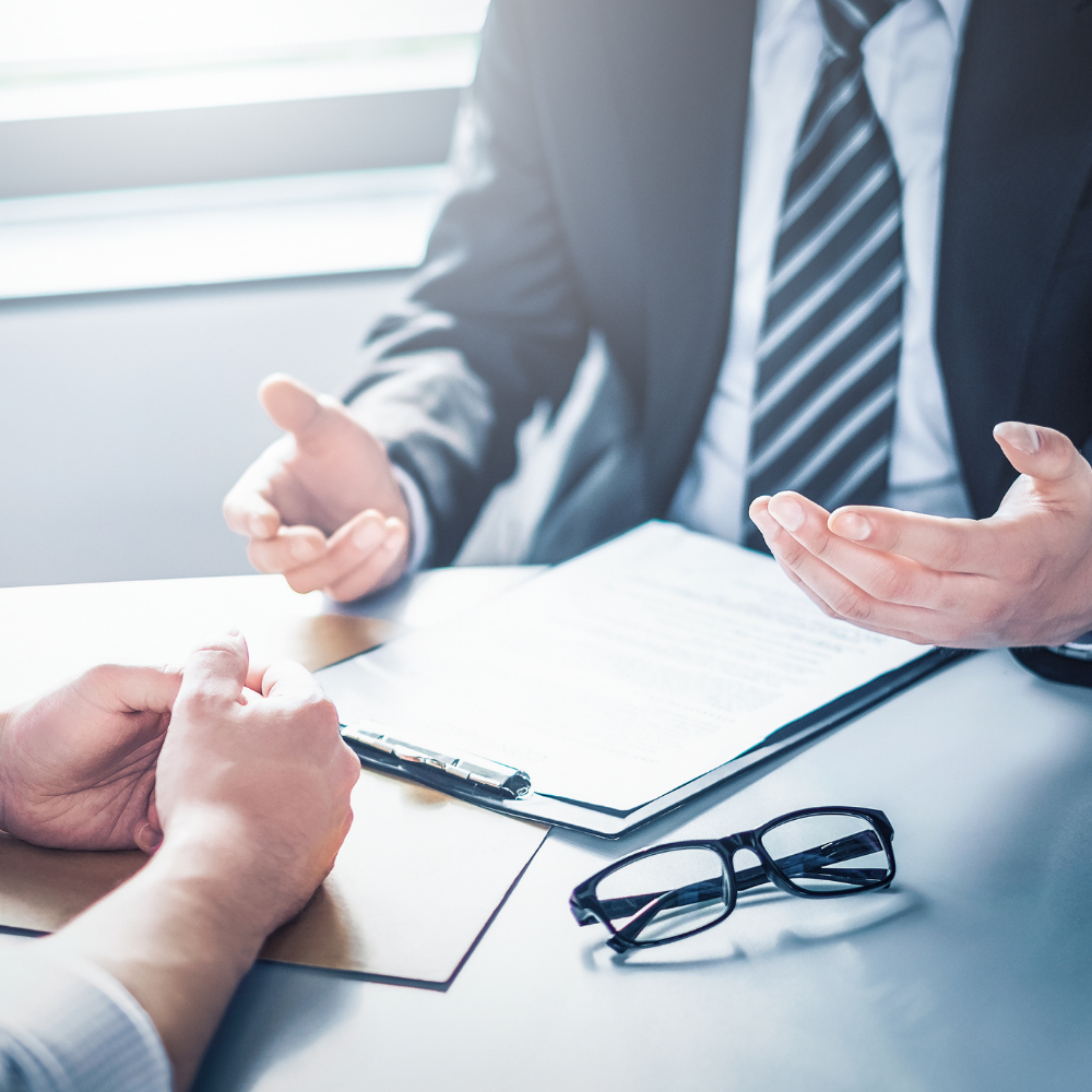 Two people at a desk, one gesturing while the other listens, paperwork, glasses.