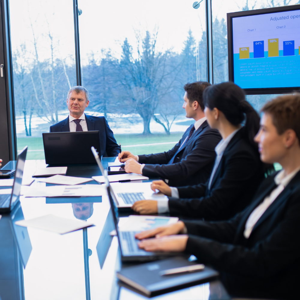 Business team in a meeting, around a table with laptops, looking at presentation on screen, bright room.