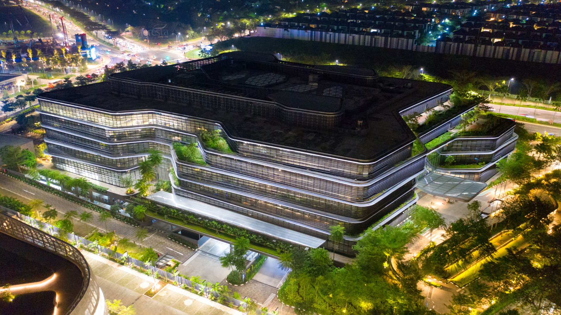 Nighttime aerial view of a large modern building with multiple levels, featuring lit windows and greenery, with city lights in the background.