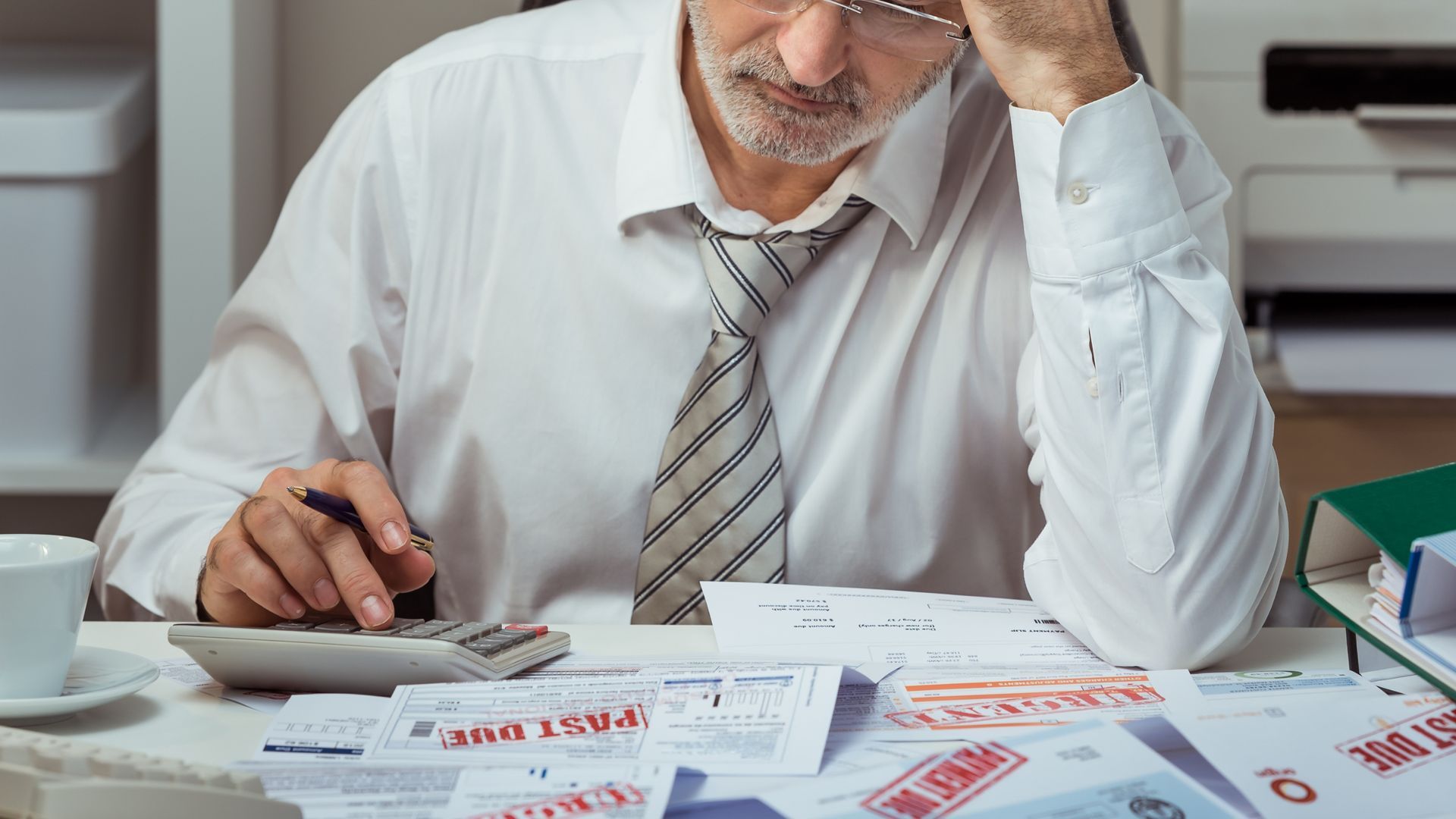 Man stressed, calculating finances with a calculator, surrounded by bills at a desk.