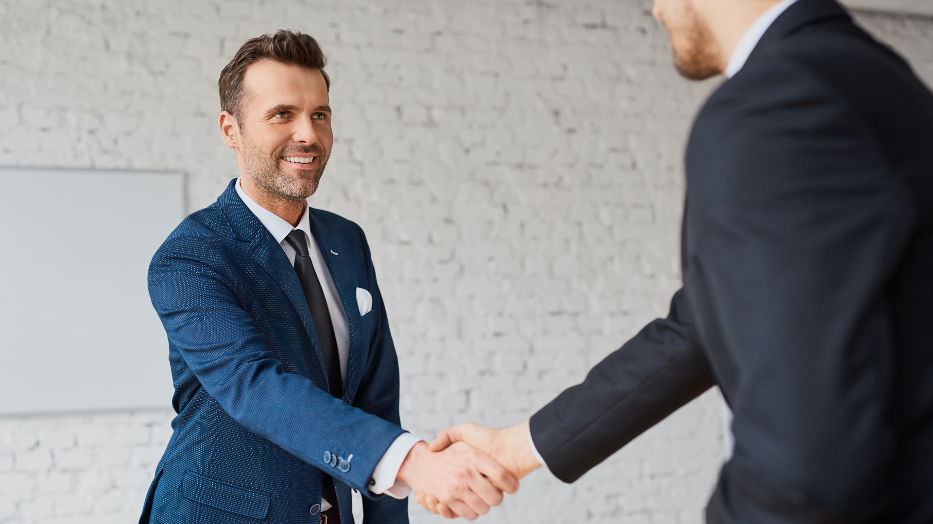 Two men shaking hands in an office; one smiling, wearing a gray blazer and blue shirt.