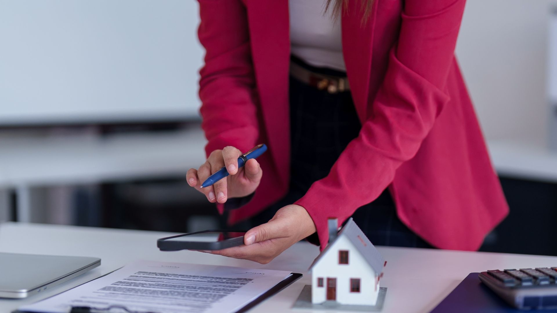 Person in red blazer holding a pen and phone, looking at a miniature house on a desk with documents.