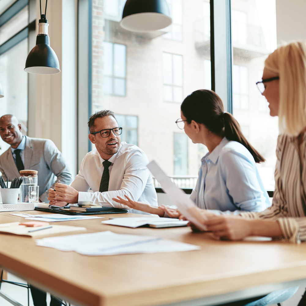 Business meeting around a table. People talking and reviewing documents. Modern office with large windows.