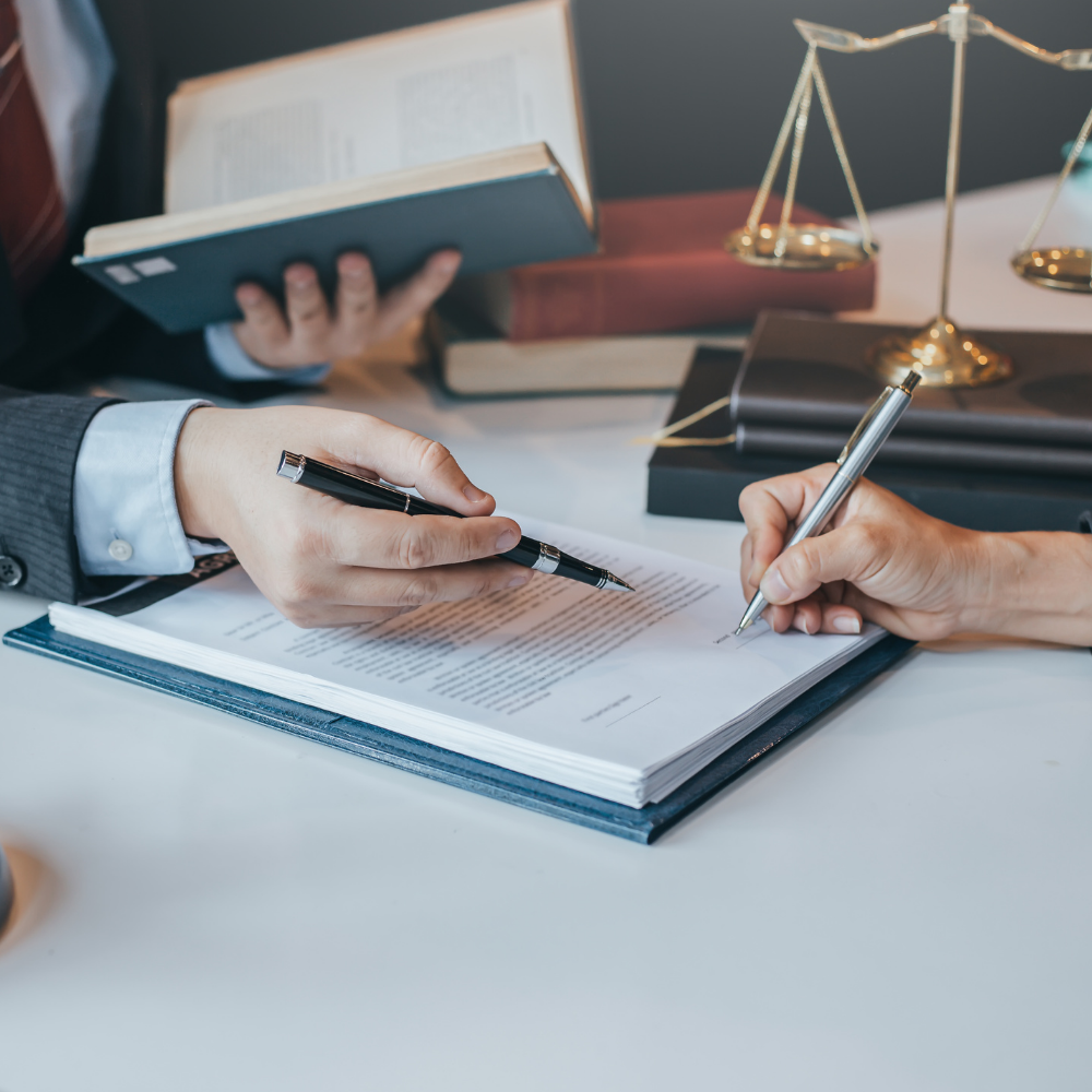 Person signing legal documents with another person holding a pen, scales of justice in the background.