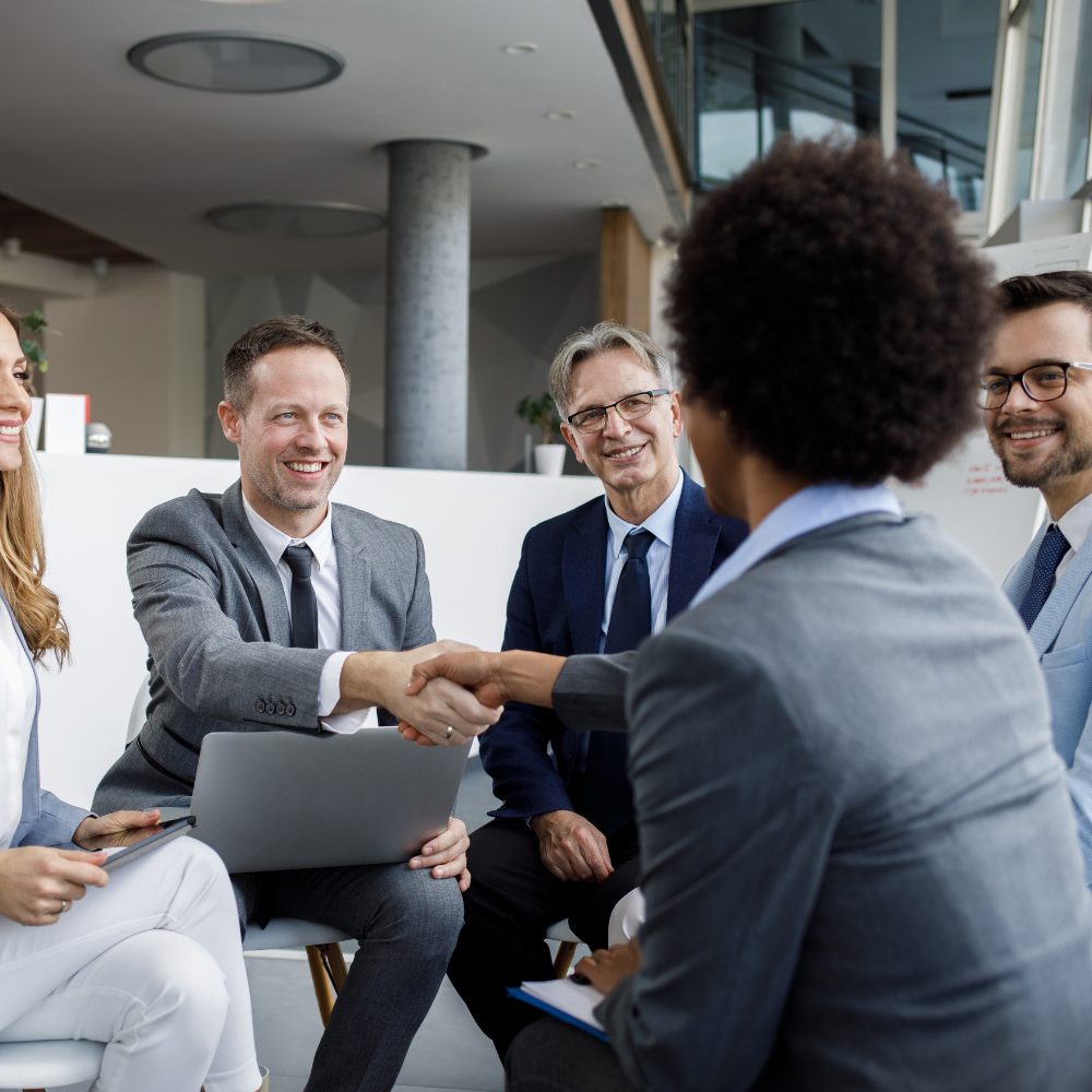 Businesspeople in suits shake hands while seated, smiling. Laptop and pad visible.