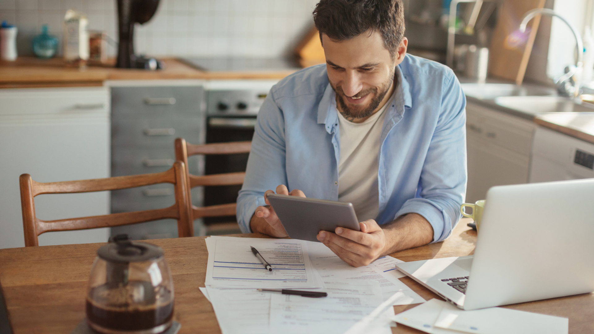 Man smiling, looking at a tablet, working at a kitchen table with a laptop, bills, and coffee.