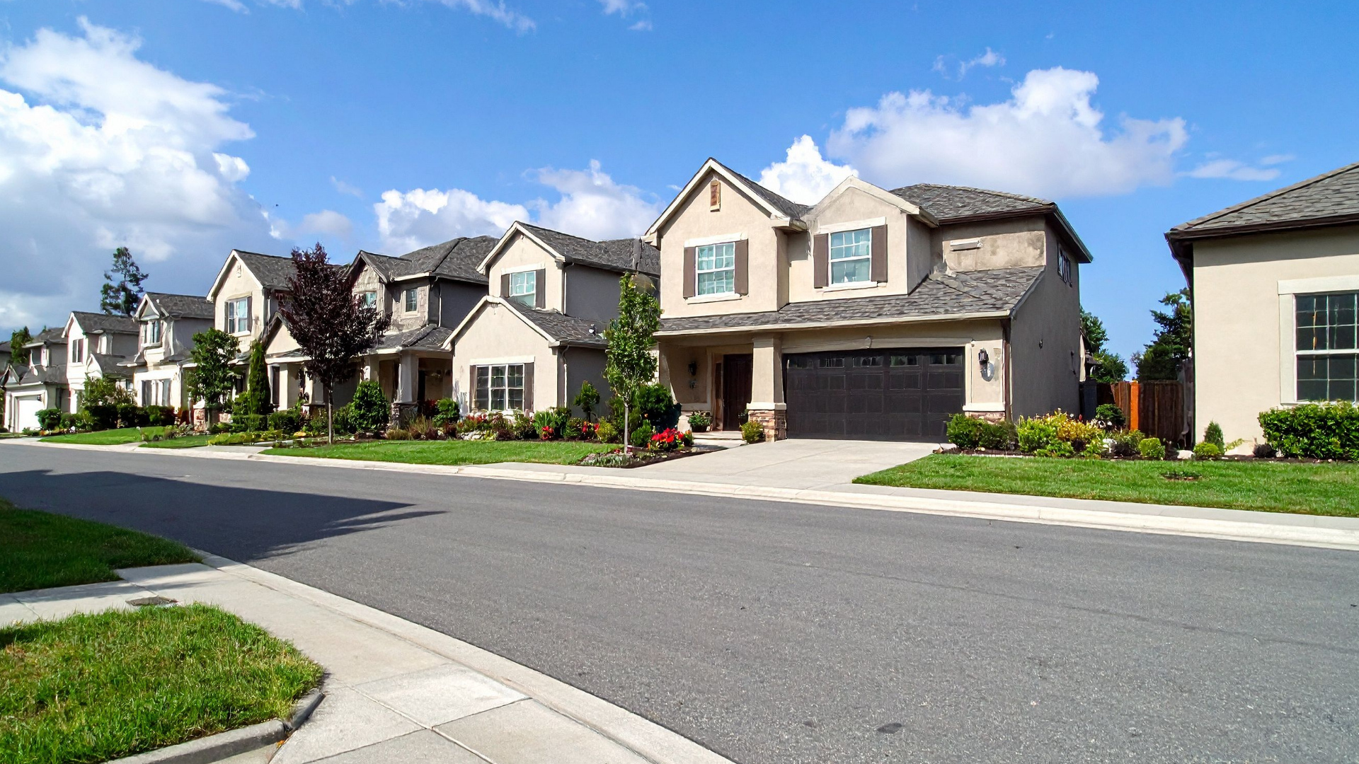 Row of houses with tan exteriors, grey roofs, and a paved street on a sunny day.