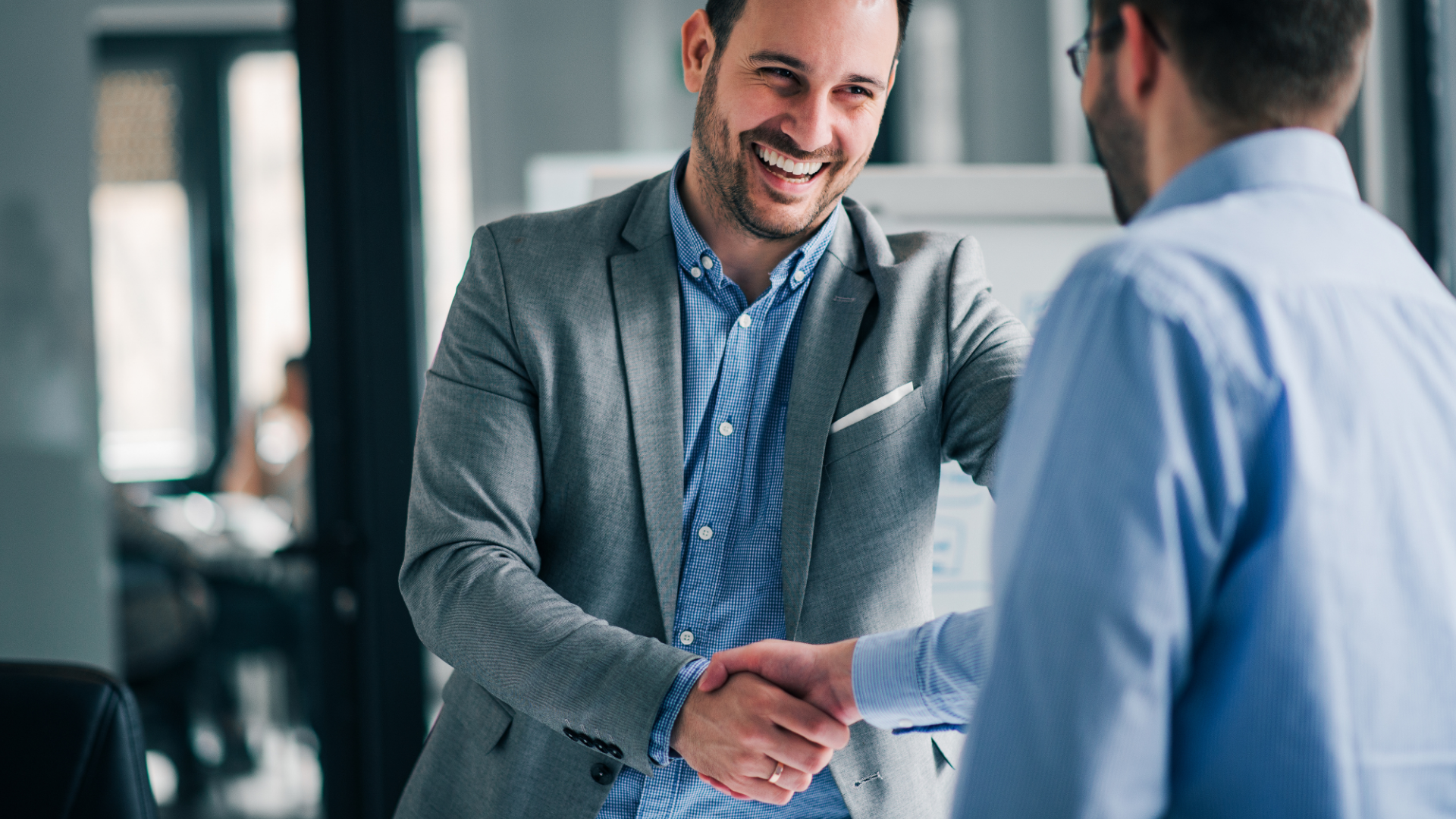 Two men shaking hands in an office; one smiling, wearing a gray blazer and blue shirt.