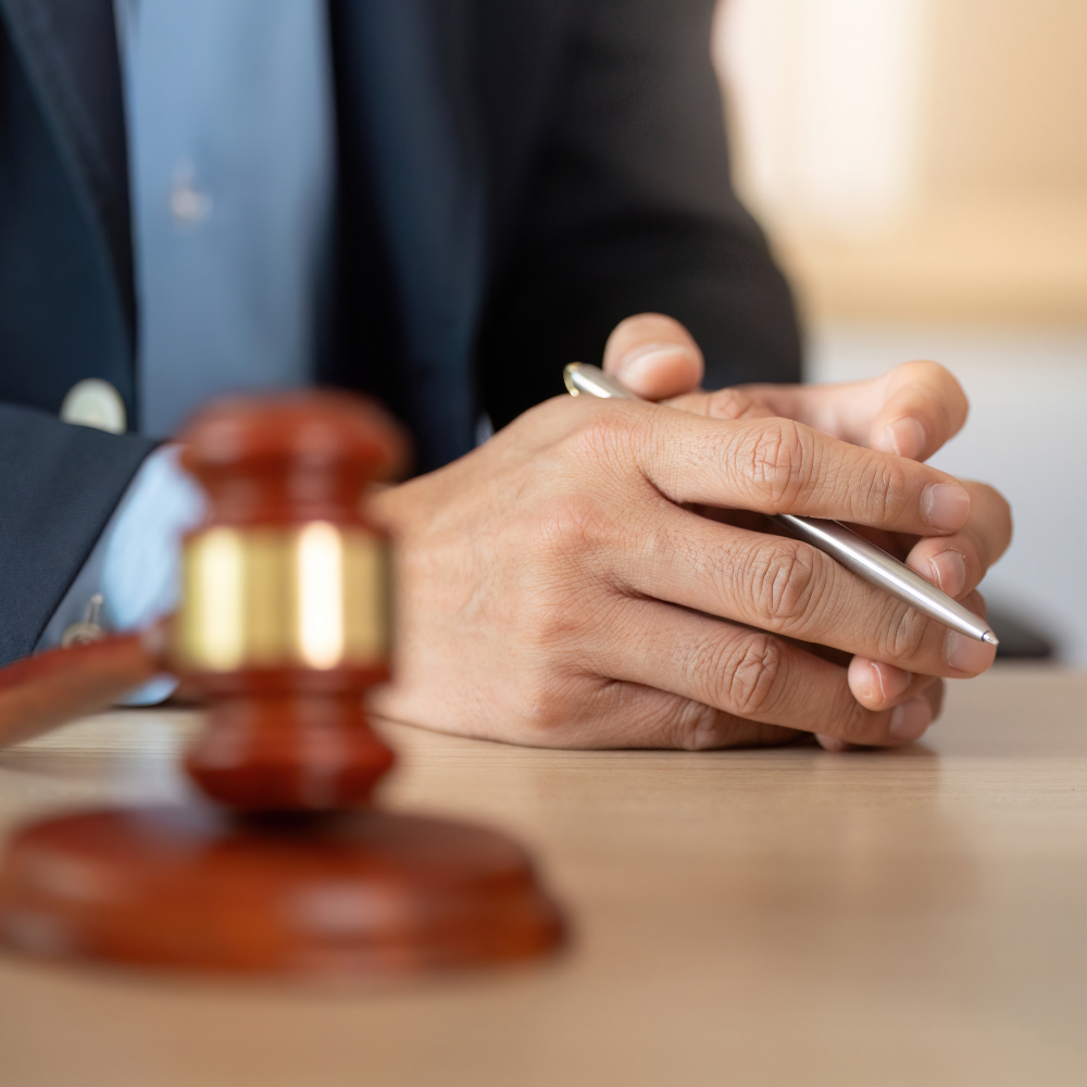 Wooden gavel and person in suit with clasped hands holding a pen.