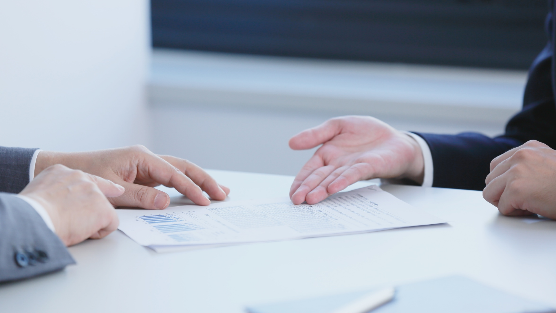 Two people in suits at a table reviewing documents, one gesturing towards the paper.