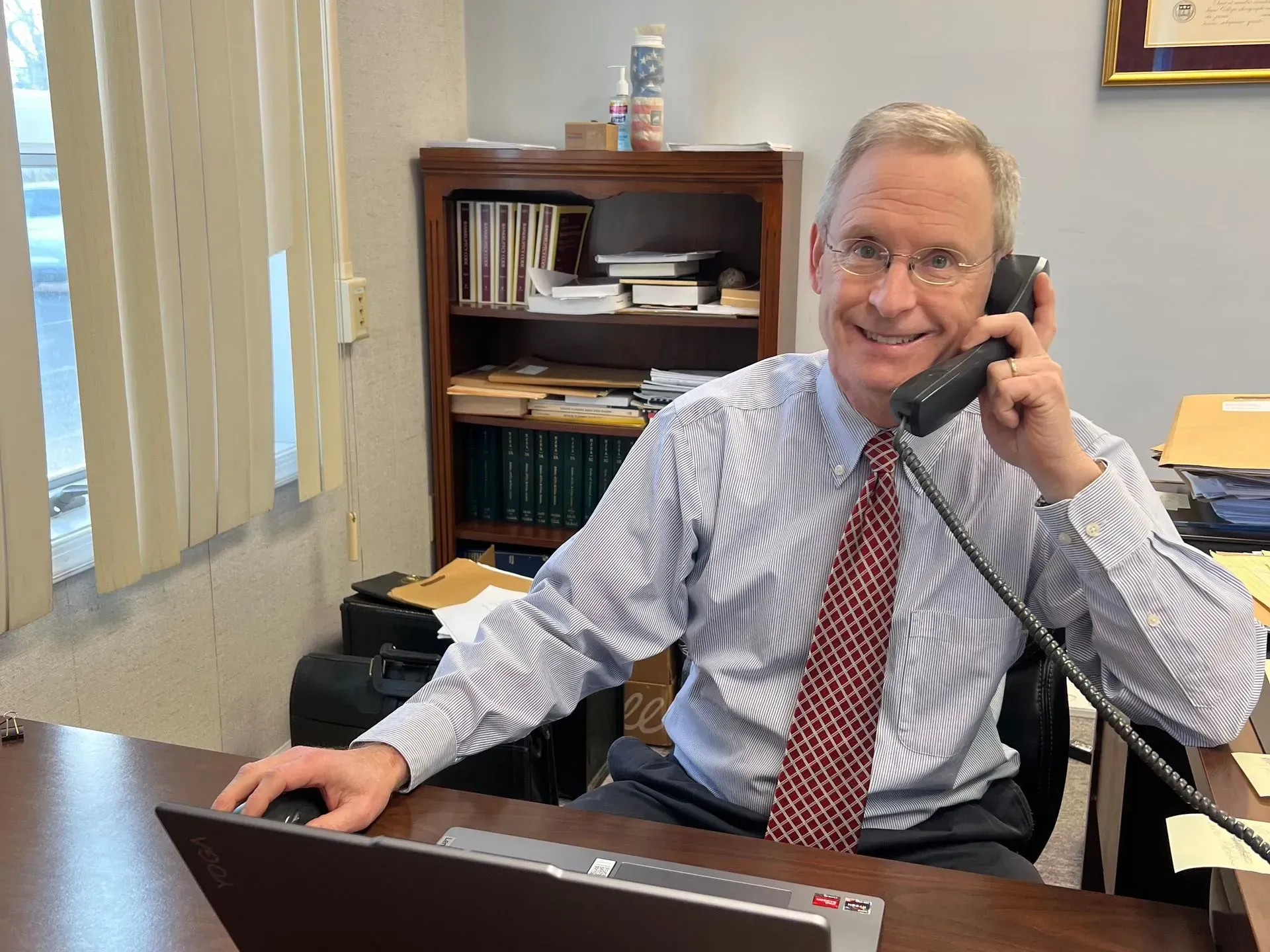 Man in office smiles, holding phone, seated at desk with computer. Bookshelf in background.