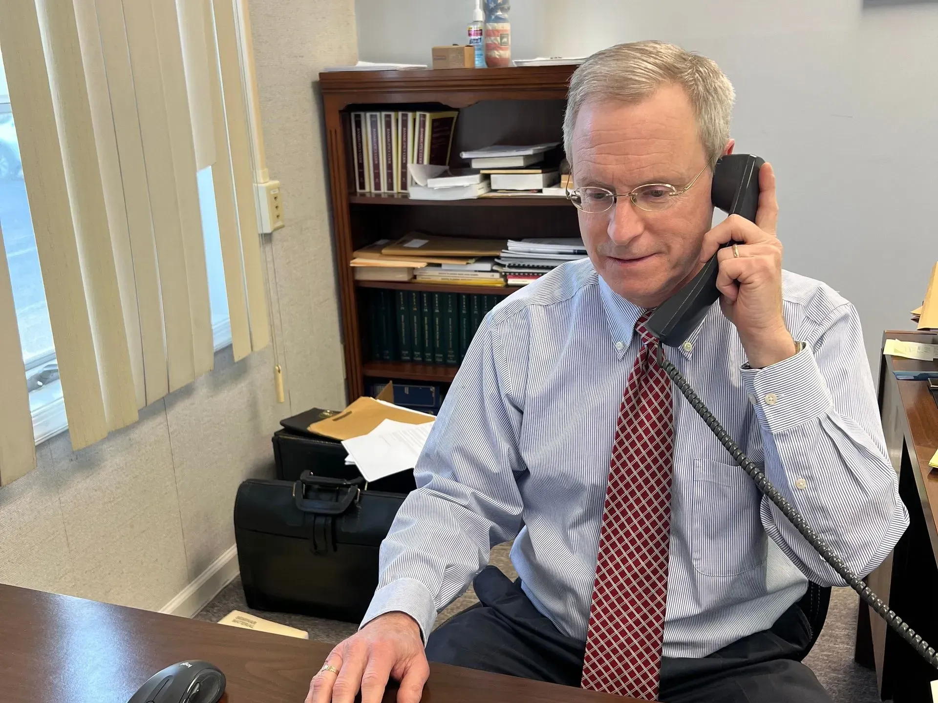 Man in office on phone, wearing glasses and tie, sitting at desk. Bookshelf in background.
