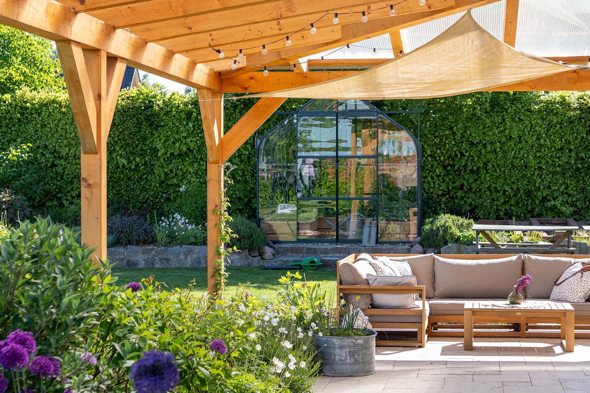 Wooden pergola over a patio with outdoor seating, a greenhouse, and lush green garden.