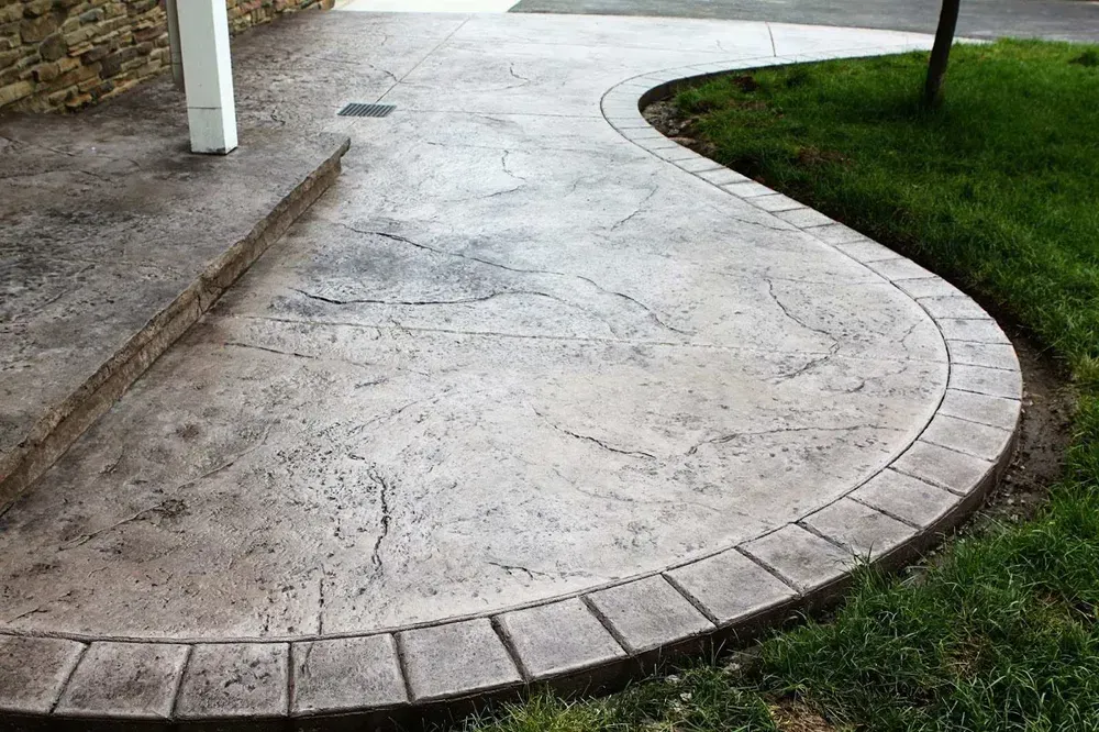 Curved gray concrete patio with a brick-like border, adjacent to green grass and a stone wall.