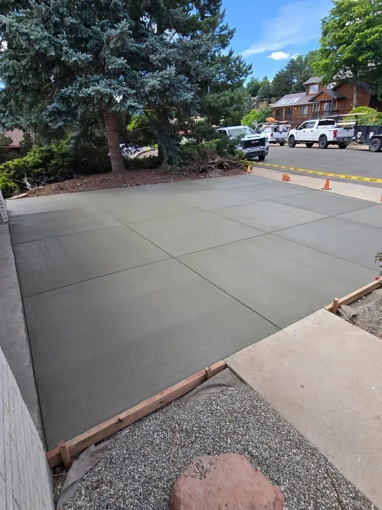 Freshly poured concrete driveway with cut lines, in front of a house with trees and parked vehicles.