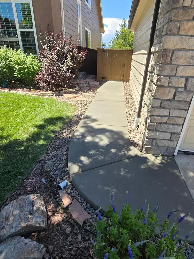 Concrete walkway alongside a house, leading to a wooden gate. Green lawn and bushes are on the left.