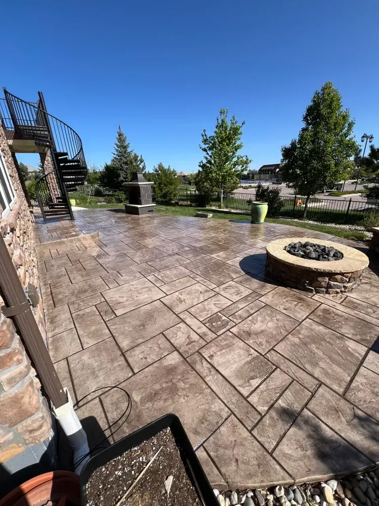 Stamped concrete patio with a fire pit, stone accents, spiral staircase, and green plants under a blue sky.