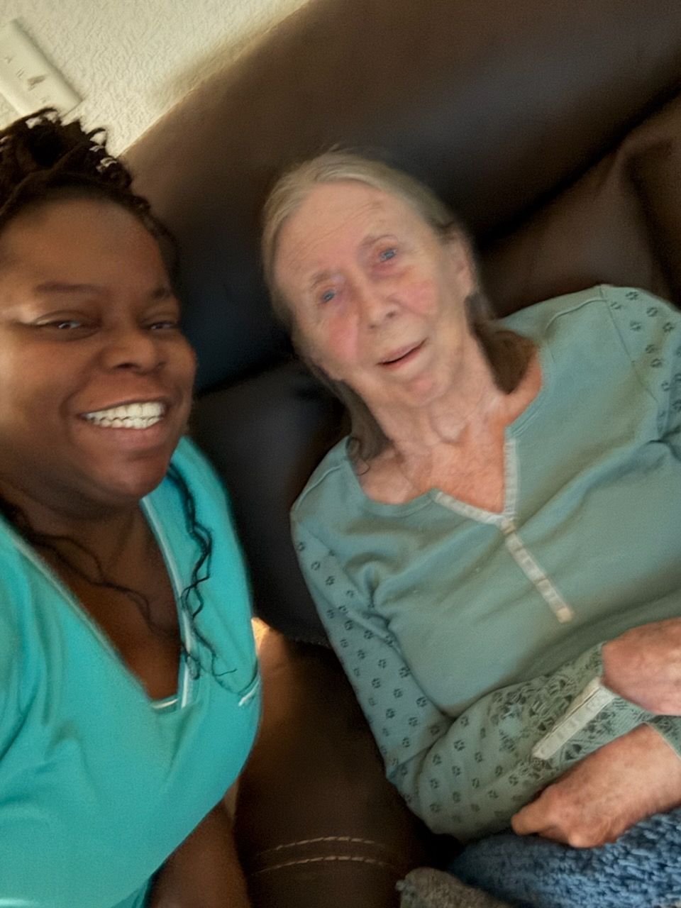 Woman smiles next to older woman on a dark couch. Both wear blue tops, indoors.