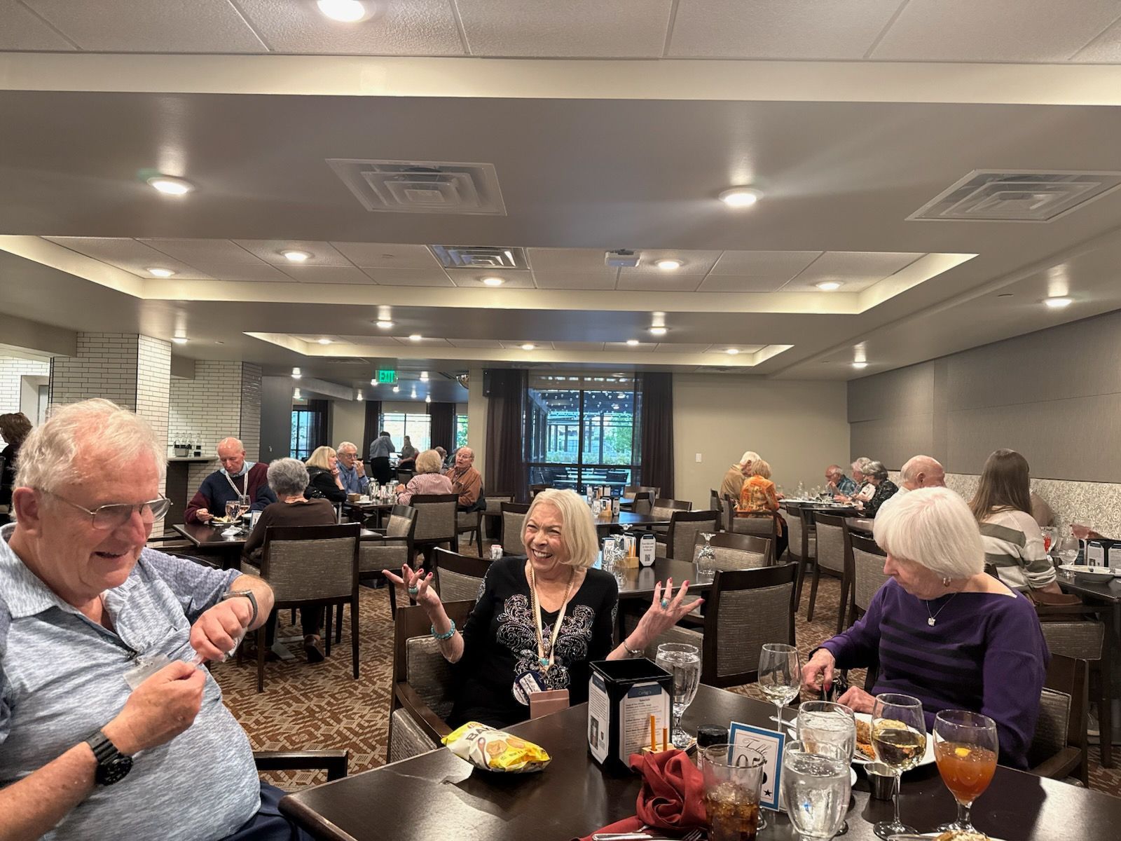 People seated at tables in a restaurant; some are eating, smiling, and talking. Brown tables and chairs.