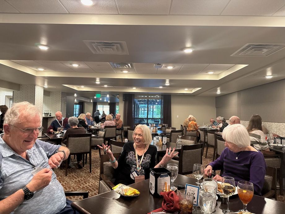 People seated at tables in a restaurant; some are eating, smiling, and talking. Brown tables and chairs.